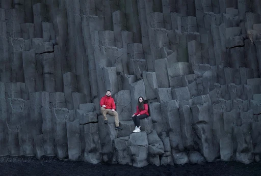 Couple in red jackets sitting on dramatic basalt columns at Iceland's Reynisfjara Beach.