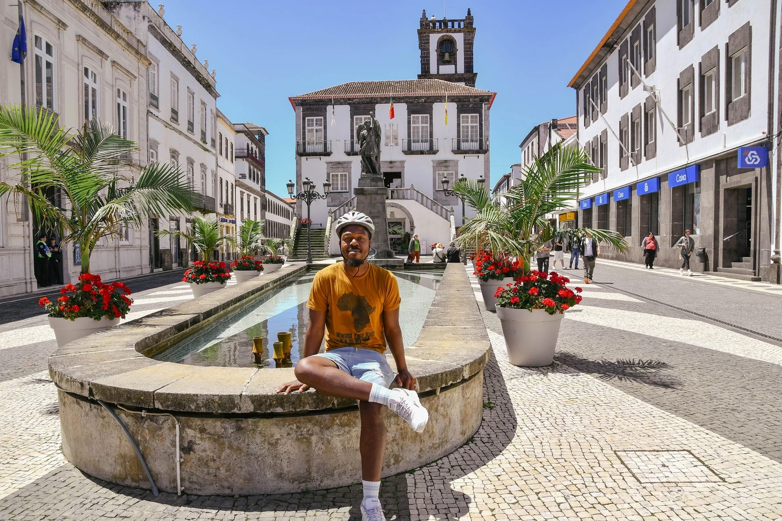 A man sits by a fountain in Ponta Delgada, Azores, under bright daylight, surrounded by historic architecture.