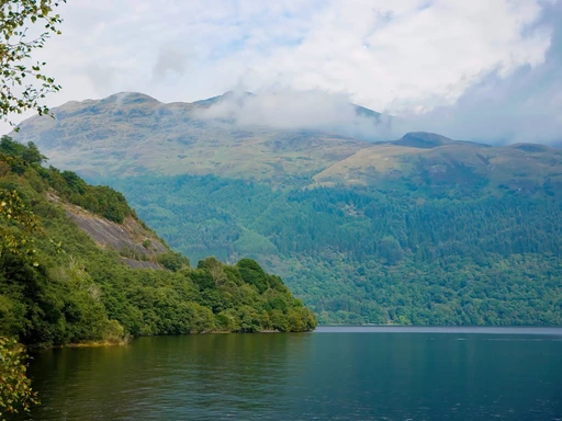 Beautiful landscape of Loch Lomond with lush green hills and misty mountains in the background.