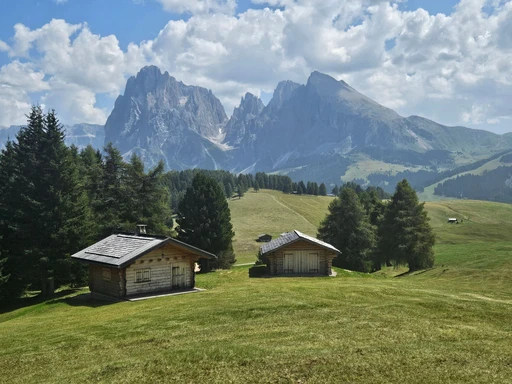 Two wooden huts on a grassy slope with a Dolomite massif behind.