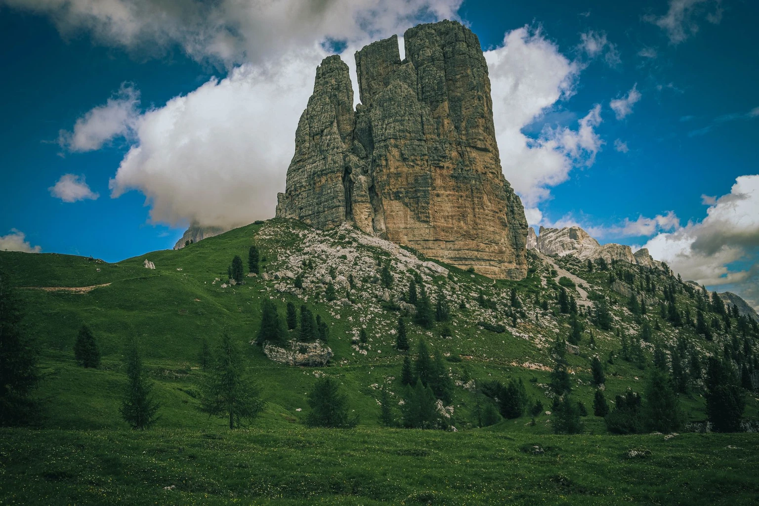 The tallest of the Cinque Torri rock towers above green slopes and pine trees.