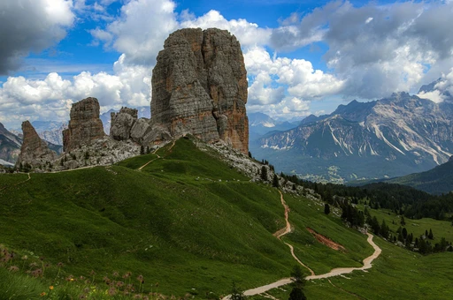 A Cinque Torri tower beside a stone-paved path with mountains under cloud behind.