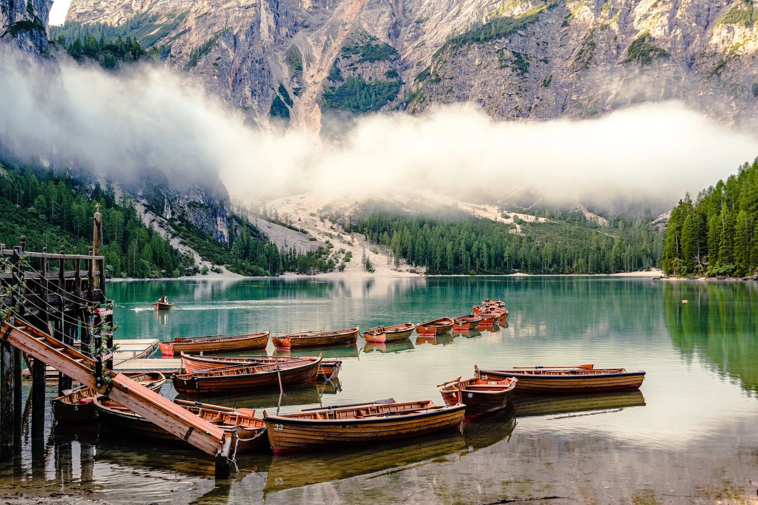 Wooden rowing boats moored on Lago di Braies with mist and cliffs behind.