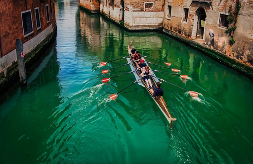 A crew rowing a long boat along a green canal in Venice, seen from a bridge.