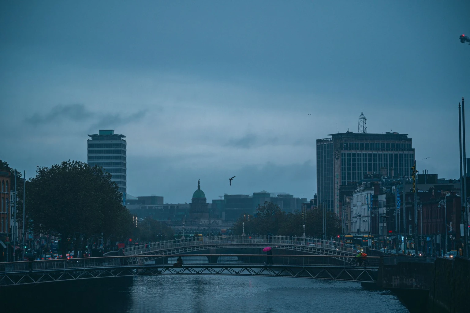 Moody Dublin cityscape showcasing Ha'penny Bridge over the Liffey River at dusk with an overcast sky.