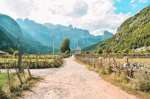 A dirt road through Theth village with wooden fences, a small stone church and the peaks of the Albanian Alps behind.