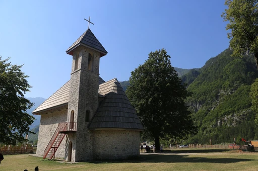 A quaint stone church in Theth, Albania, surrounded by lush mountains and trees during a sunny day.