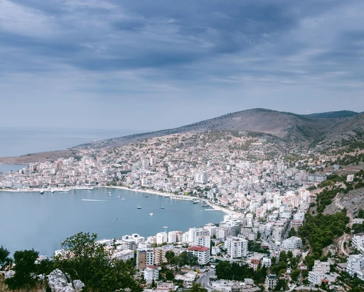 Aerial view of Sarandë on its bay, with hills behind and a marina along the waterfront under a cloudy sky.