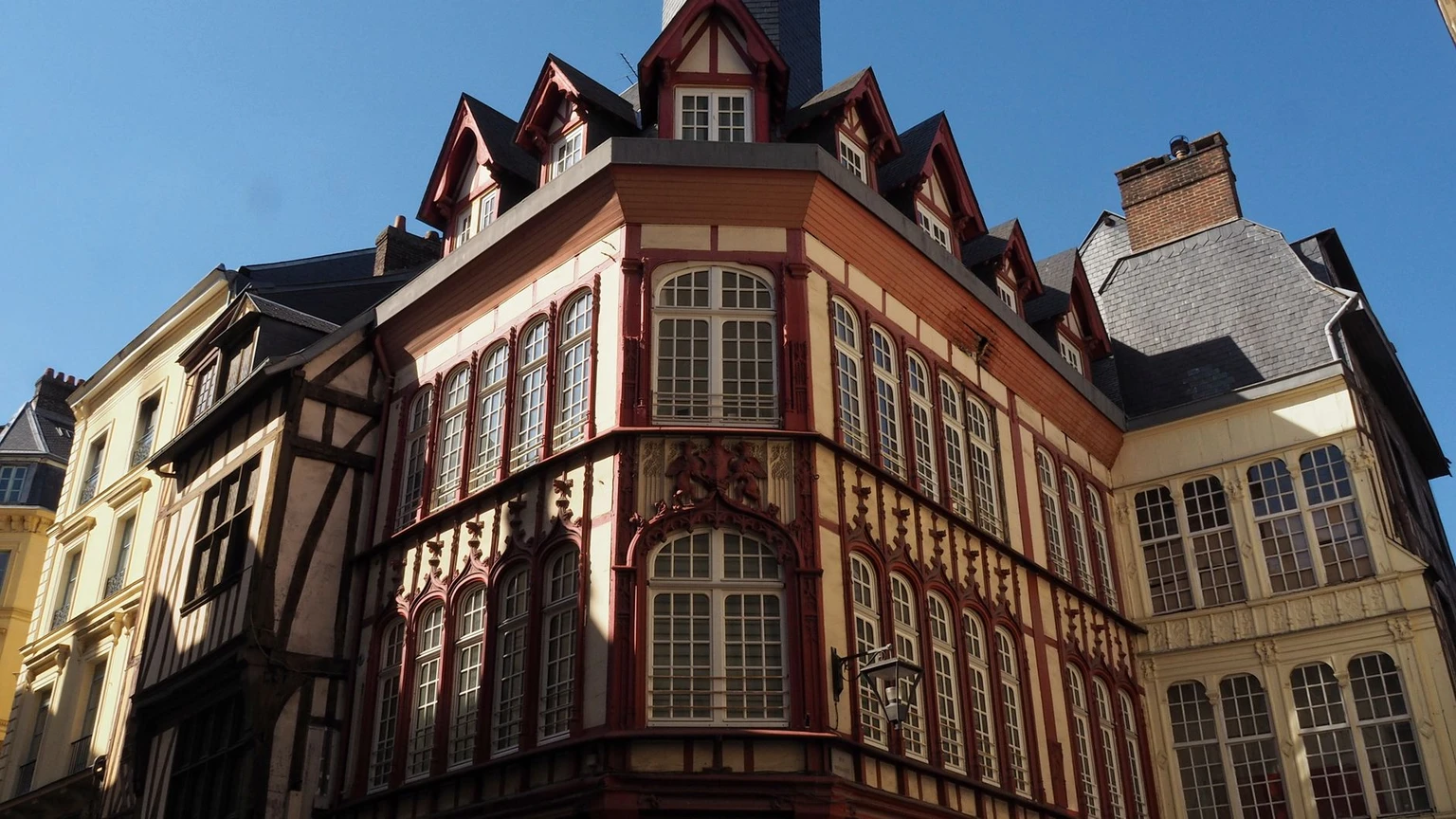A corner house in Rouen with a half-timbered upper story and tall windows, in sunshine.