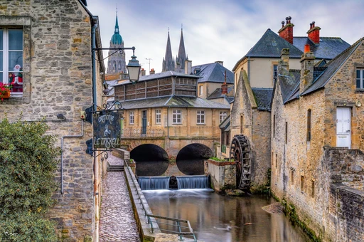 A stone watermill over a canal in Bayeux, with cathedral spires rising behind the rooftops.
