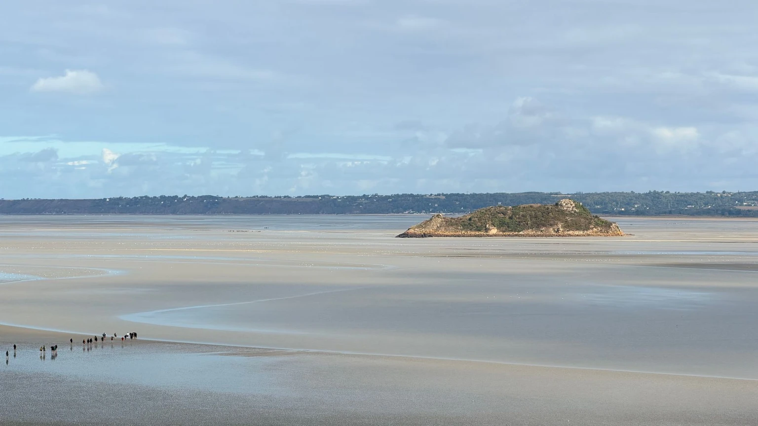 Walkers on the wet sand of the bay at low tide, with the islet of Tombelaine in the distance.