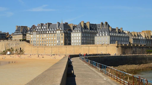 The granite walls and tall houses of Saint-Malo above the beach and a stone causeway, under a blue sky.