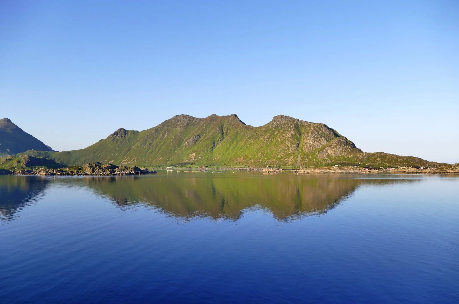 Green Lofoten mountains reflected in calm blue sea under a clear summer sky. Stand-in for the wall of mountains seen from the ferry; exact place unconfirmed.