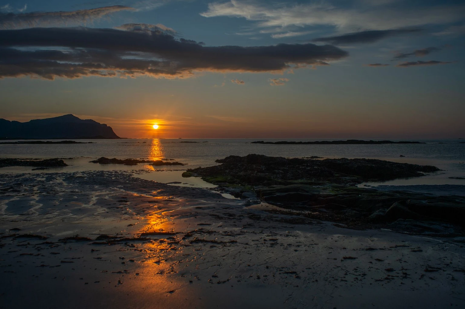 The low sun touching the sea beyond a tidal beach in Nordland, Norway. Stand-in for the midnight sun; exact beach unconfirmed.
