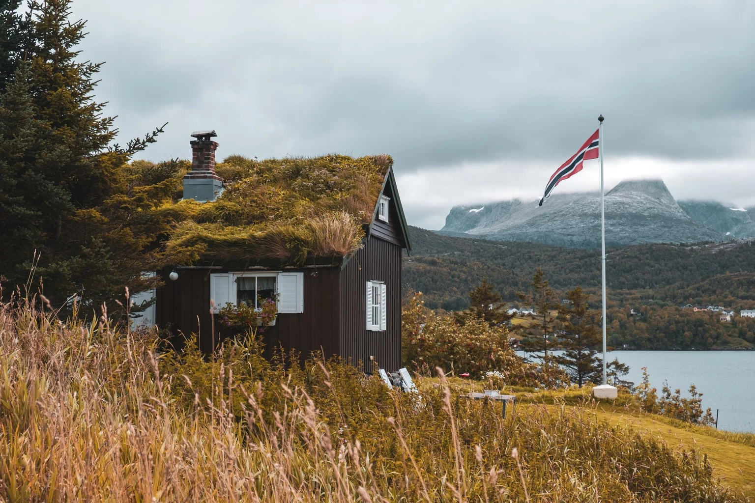 A grass-roofed wooden cottage by a fjord near Bodø, with the Norwegian flag and mountains beyond, at Saltstraumen.