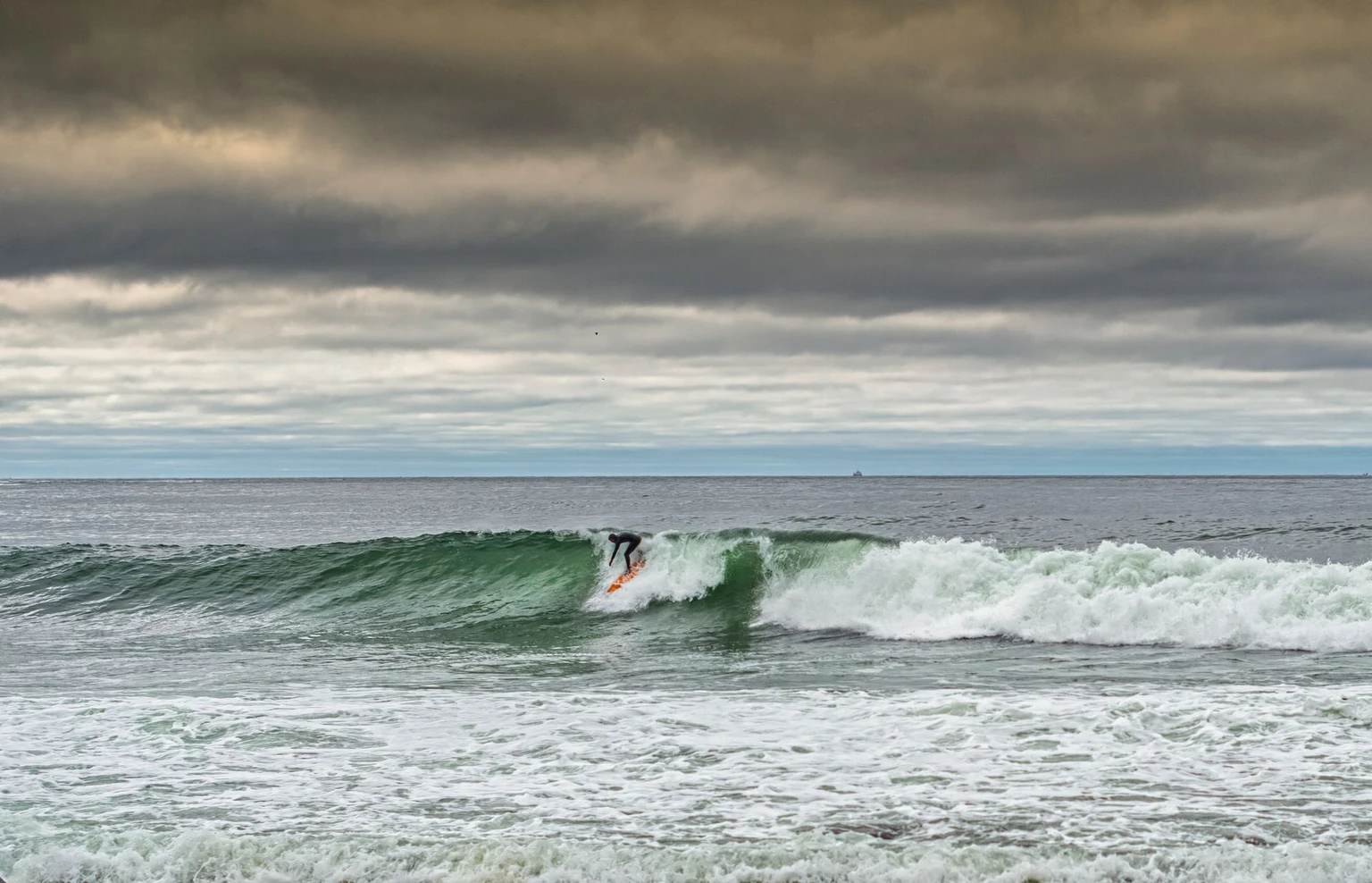 A surfer riding a green wave under heavy cloud at Unstad beach, Lofoten.
