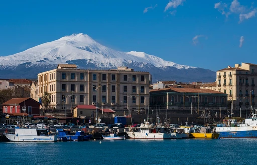 Snow-capped Mount Etna above the fishing boats and waterfront buildings of Catania's harbor.