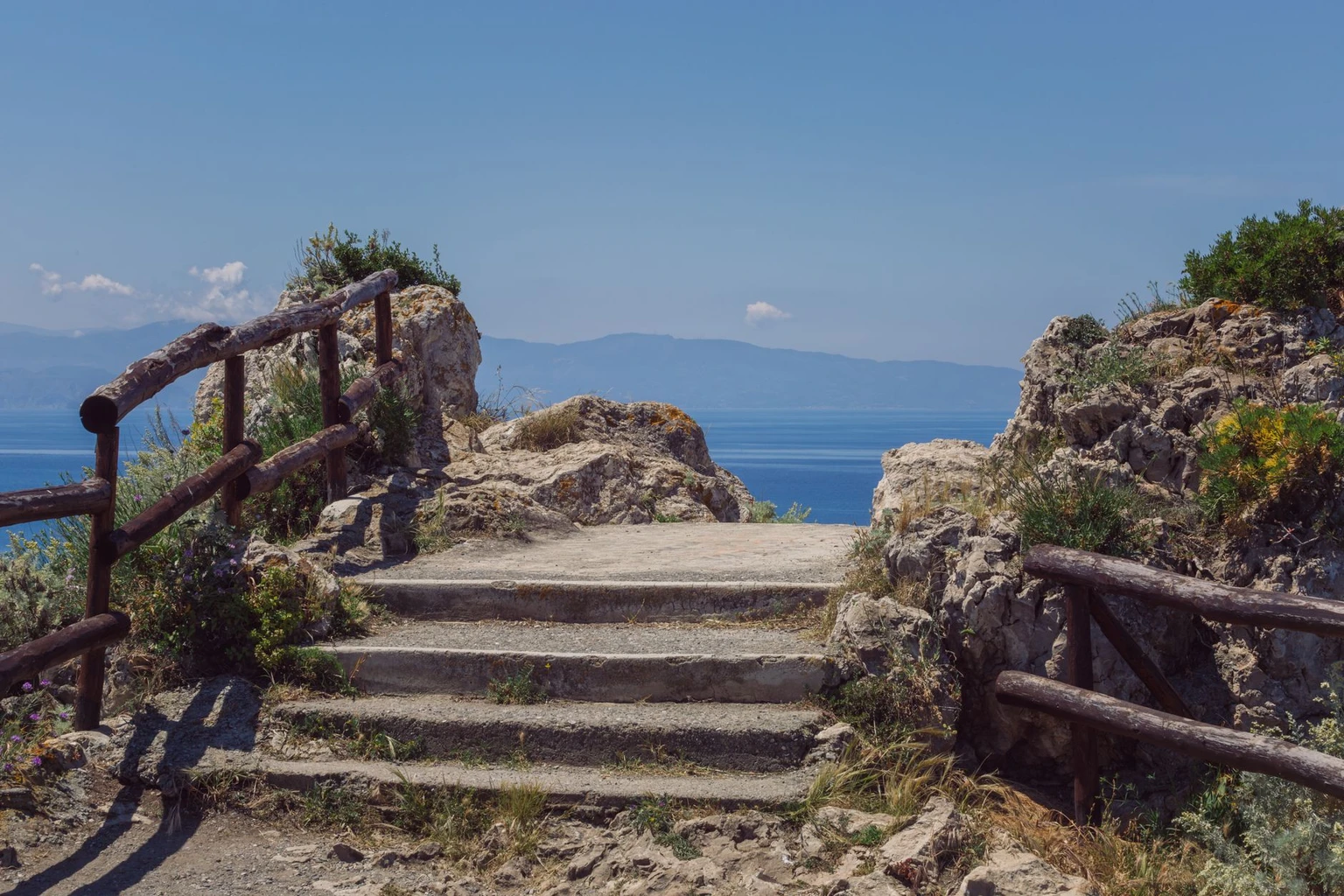 A stone path with steps and wooden railings leading to a cliff-top viewpoint over the sea at Milazzo, Sicily.