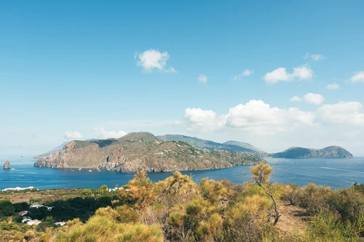 A rocky headland with a railed path above the sea, with other Aeolian islands on the hazy horizon.