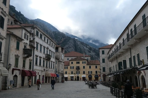 Kotor's old-town square on an overcast day, with a few people crossing it and cloud over the mountain behind.