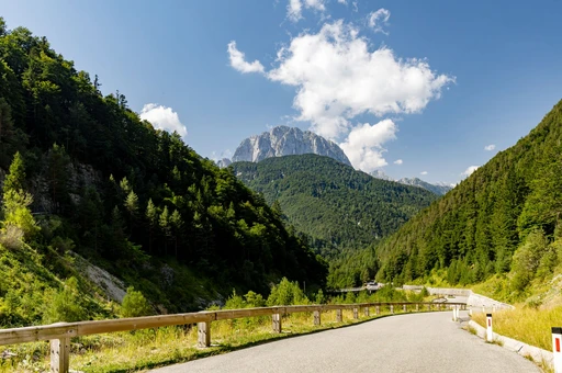 A panoramic view of a winding road through lush green forests and the majestic Julian Alps under a clear blue sky.