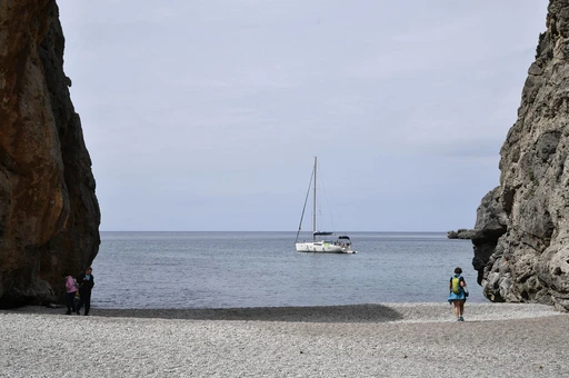 The pebble beach at the mouth of the Torrent de Pareis between cliff walls, with a sailboat anchored beyond.