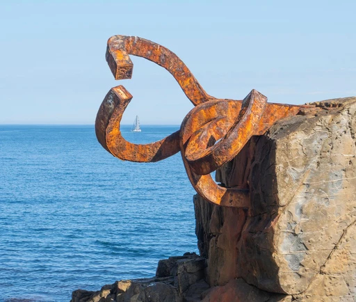 A rust-colored steel sculpture of Chillida's Peine del Viento on the rocks at the end of San Sebastián's bay, with a sailboat on the sea beyond.