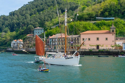 A traditional sailboat with a red sail passing moored boats beside a pink stone building in a Basque harbor, believed to be the Pasaia channel.