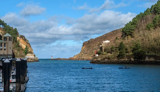 Two rowing boats on calm water in a channel between green cliffs, with a small lighthouse at the far end, believed to be the mouth of the Pasaia channel.
