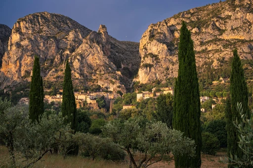 Moustiers-Sainte-Marie under its limestone cliffs at golden hour, with cypress trees in front.