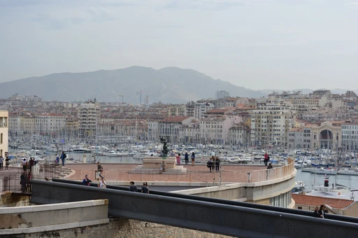 Marseille's Vieux-Port and city skyline seen from a viewpoint, with people walking along the quay.
