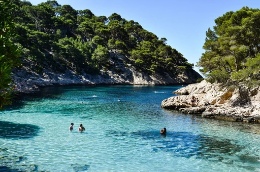 A calanque cove with two swimmers wading in shallow turquoise water beside rocks.