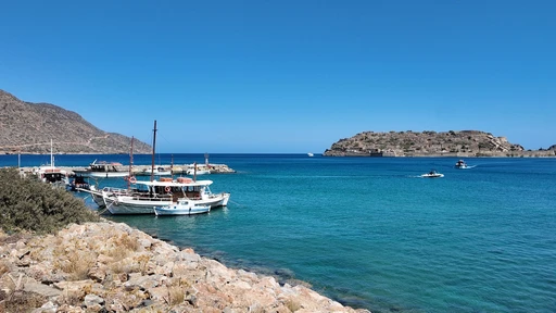 Fishing boats moored on a rocky shore near Plaka, with Spinalonga island low on the horizon.