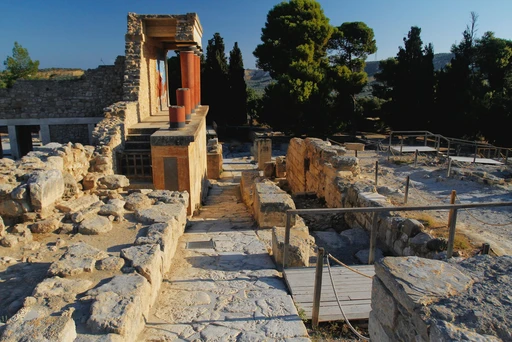 Stone steps and ruins of the palace at Knossos, with a red column above and pines behind.