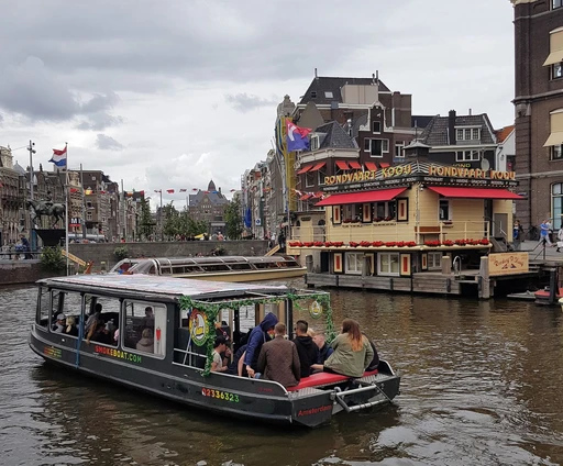 A small boat with people aboard on an Amsterdam canal, with gabled houses behind.