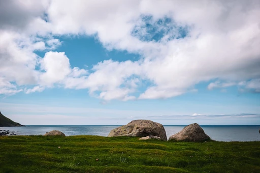 Grass and large boulders on the open shore at Eggum, Lofoten, with the sea behind under a summer sky.