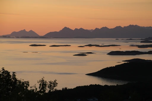 The midnight sun turning the sky orange over calm sea and low islets off Bodø, with a line of mountains on the horizon, on a July night.