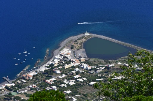 Lingua on Salina seen from high above: the village, the salt pond behind its shingle spit and the lighthouse at the tip, in deep blue sea.