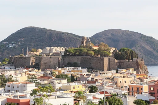 The castle walls of Lipari on their rock above the town, with the cathedral bell tower behind them and two green hills beyond.